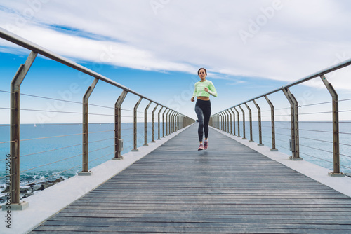 Young female jogger in active clothes reaching running goals on cardio session during morning time, slim Caucasian sportswoman training athletic strength enjoying workout at boardwalk pier