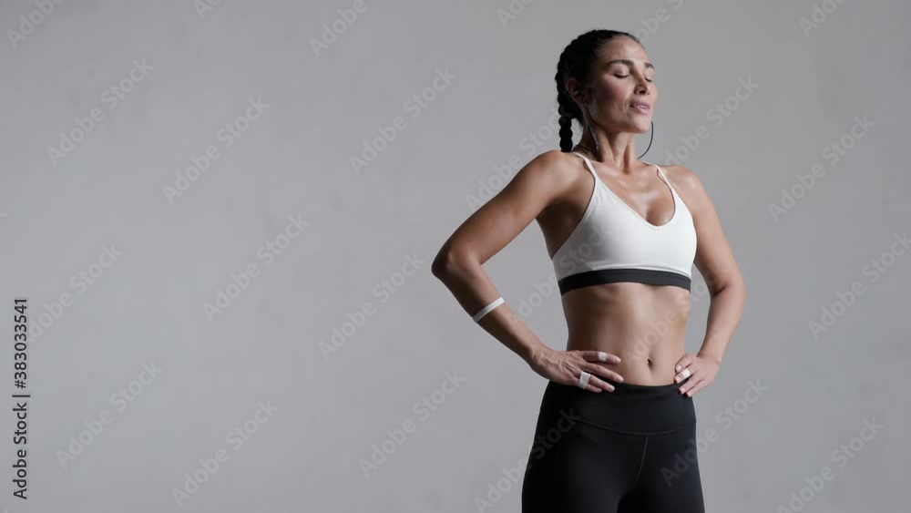 Portrait of beautiful fitness woman smiling and looking at camera isolated on grey background. Laughing mid woman in sportswear wearing wireless headphones and relaxing after training at gym.