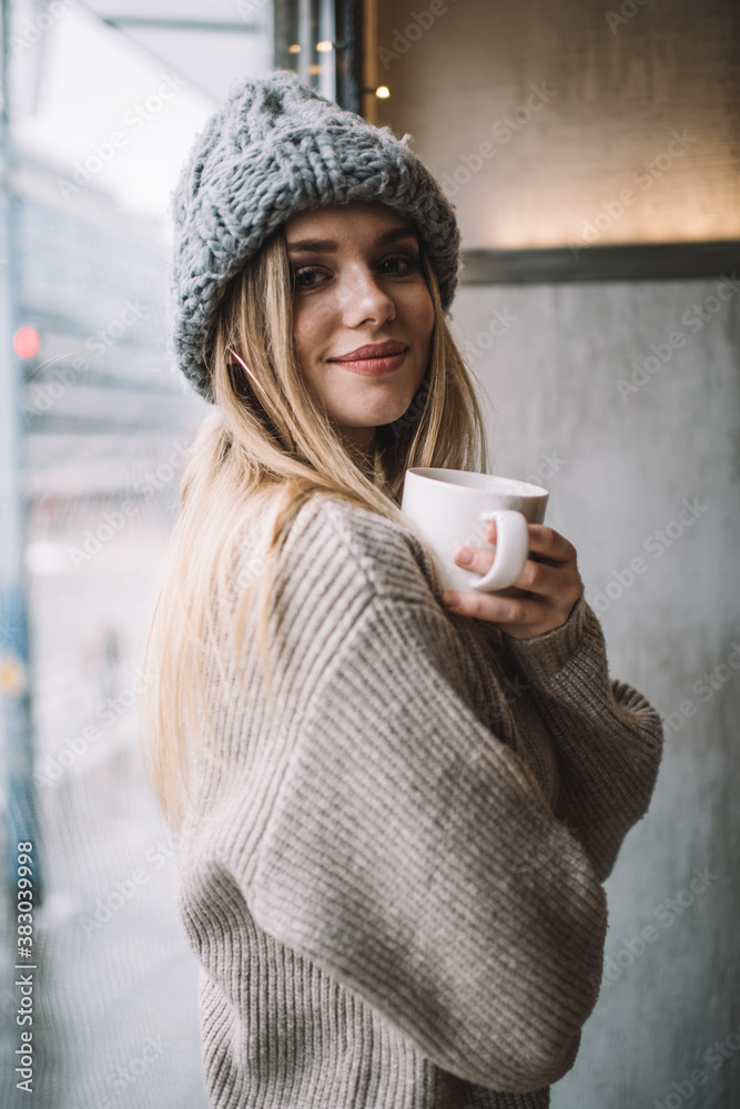 Half length portrait of trendy dressed customer posing near cafeteria window enjoying coffee time, carefree female in knitted pullover and winter hat holding aroma caffeine beverage for warming - obrazy, fototapety, plakaty 