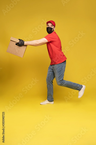 Delivery man in protective mask and gloves with box in studio isolated on yellow background