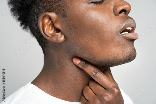 Closeup of afro man touches fingers of sore throat, isolated on gray background. Thyroid gland, painful swallowing, tonsillitis, laryngeal swelling concept. Inflammation of the upper respiratory tract photo