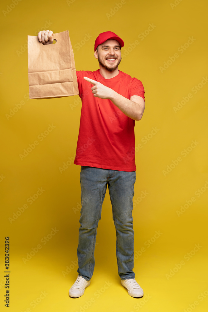 Man in red cap, t-shirt giving fast food order isolated on yellow background. Male employee courier hold empty paper packet with food. Products delivery from shop or restaurant to home. Copy space