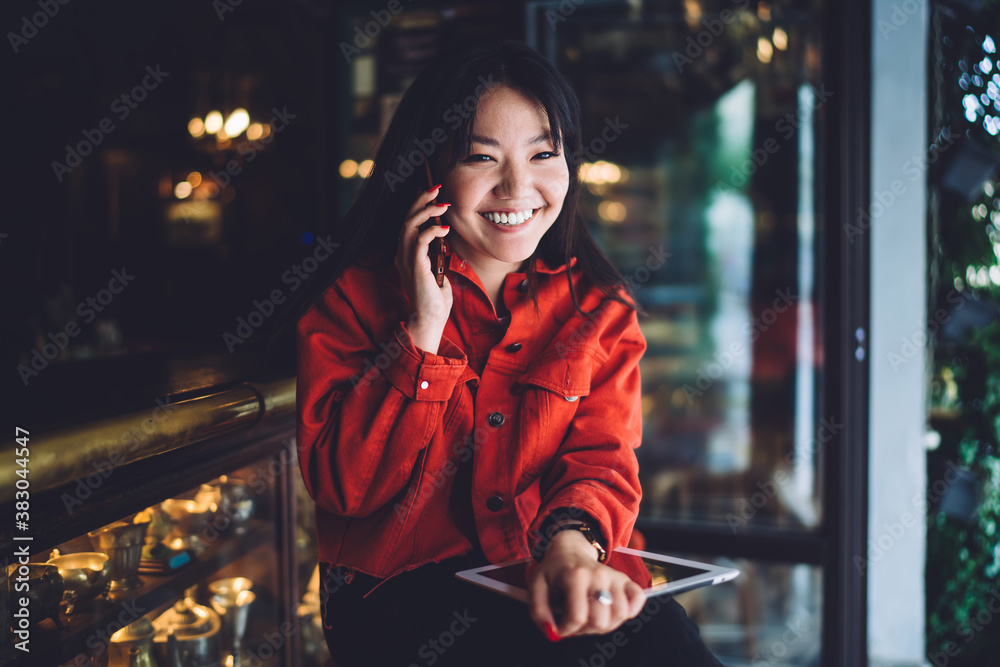 Cheerful young Asian woman with tablet talking on smartphone in cafe Cheerful young Asian woman with tablet talking on smartphone in cafe