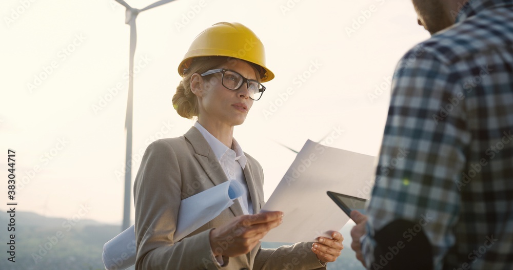 Woman in a helmet, environmental observer with documents in hands standing together with a male worker of the windmill power station and giving him orders and advice while he noting on the tablet. Woman in a helmet, environmental observer with documents in hands standing together with a male worker of the windmill power station and giving him orders and advice while he noting on the tablet.
