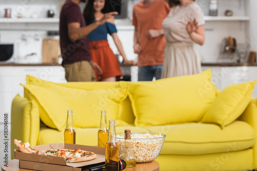 selective focus of pizza and beer on table near yellow sofa and multicultural friends dancing on background