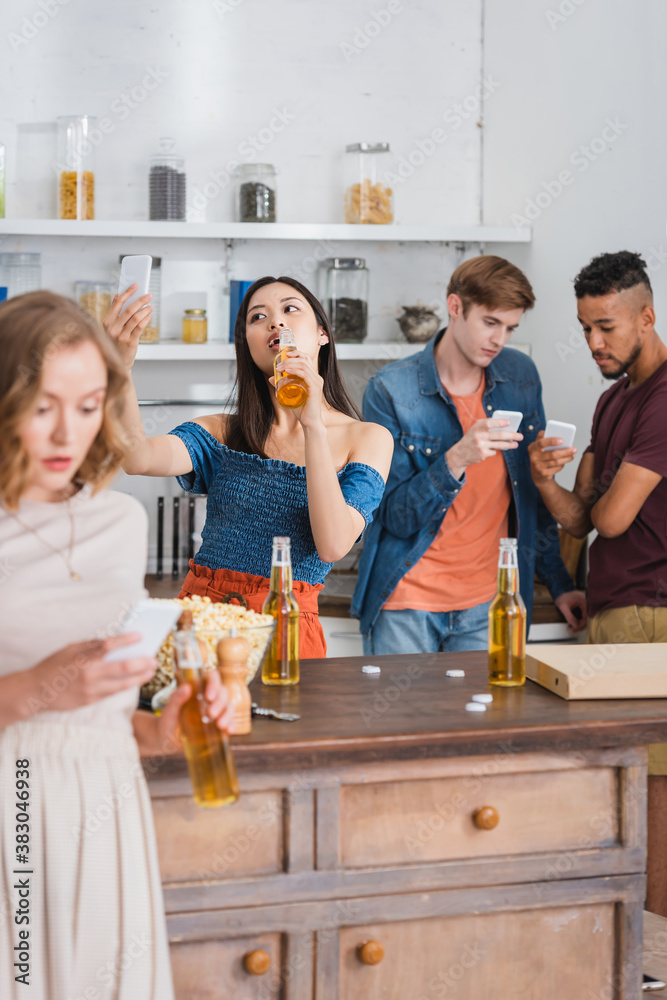 Obraz premium selective focus of asian woman drinking beer and taking selfie near multiethnic friends chatting on mobile phones during party