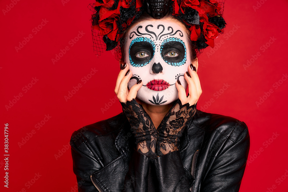 Young creepy lady calavera. wears artistic make-up for the feast of all the dead. Touches decorated face, wears black leather jacket and lace gloves, dressed as skeleton isolated in red.
