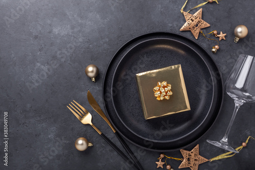 Christmas table setting with black ceramic plate, glass, gift box and gold accessories on black stone background. Top view. Copy space