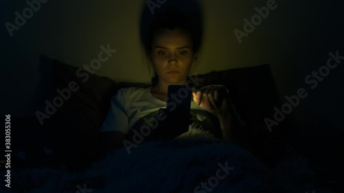 Girl using mobile phone lying in bed at night. Young woman browsing online and scrolling social media in smart phone