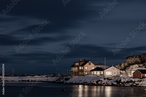 Wooden cottage on the shore of the sea at night in winter. View of harbour with wooden houses in winter season surrounded by snowy mountains