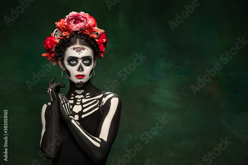 Passioned. Young girl like Santa Muerte Saint death or Sugar skull with bright make-up. Portrait isolated on dark green studio background with copyspace. Celebrating Halloween or Day of the dead.