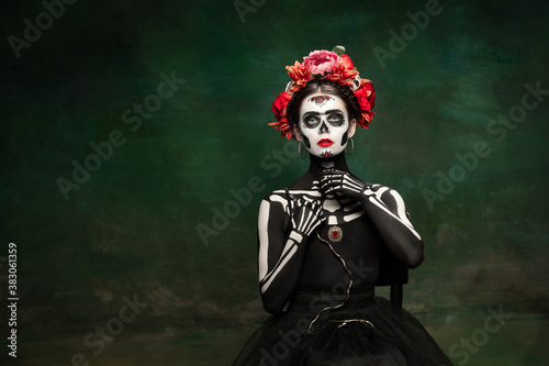 Poison. Young girl like Santa Muerte Saint death or Sugar skull with bright make-up. Portrait isolated on dark green studio background with copyspace. Celebrating Halloween or Day of the dead.