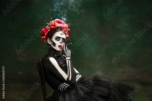 Smoking. Young girl like Santa Muerte Saint death or Sugar skull with bright make-up. Portrait isolated on dark green studio background with copyspace. Celebrating Halloween or Day of the dead.
