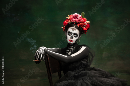 Smoke. Young girl like Santa Muerte Saint death or Sugar skull with bright make-up. Portrait isolated on dark green studio background with copyspace. Celebrating Halloween or Day of the dead.