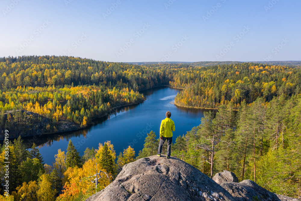The man on top of a rock in the autumn forest on a background of a beautiful lake. - obrazy, fototapety, plakaty 