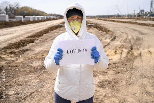 caucasian woman in protective mask stands outdoor. holds sheet of paper with the inscription COVID 19 looks at the camera. Coronavirus, health protection, quarantine, safety and pandemic concept