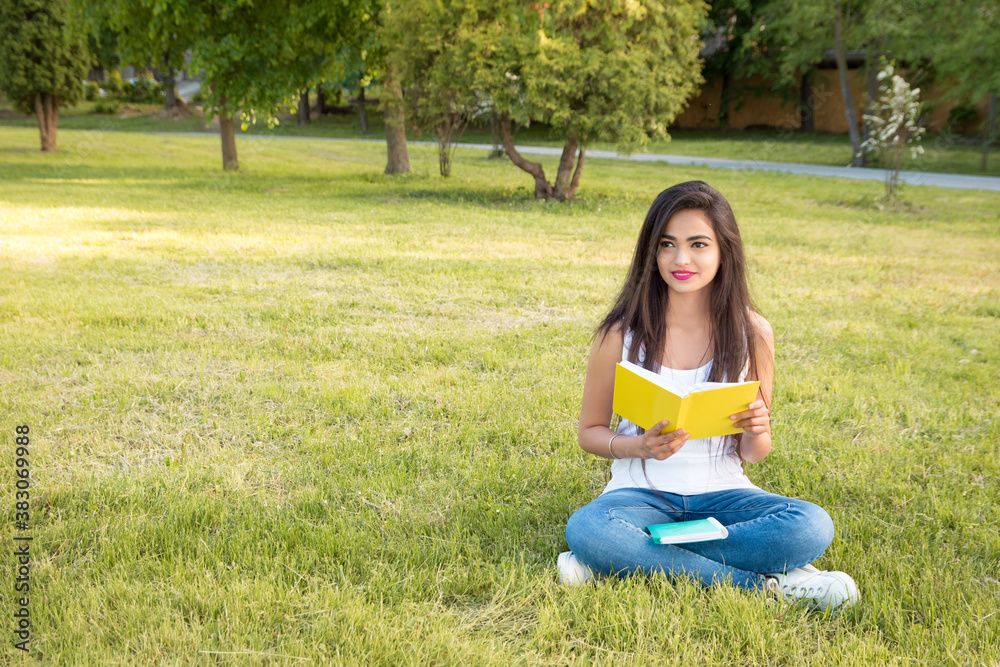 Young beautiful indian girl sitting on the grass in the park, reading a book or browsing her own notes. Portrait of a female student outdoors. She looks away Young beautiful indian girl sitting on the grass in the park, reading a book or browsing her own notes. Portrait of a female student outdoors. She looks away