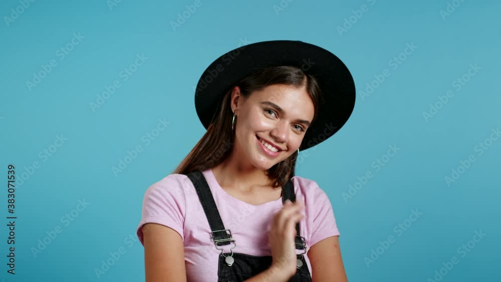Attractive hipster playful woman in overall wear and hat smiling, flirting to camera over blue wall background. Cute trendy girl's portrait Attractive hipster playful woman in overall wear and hat smiling, flirting to camera over blue wall background. Cute trendy girl's portrait