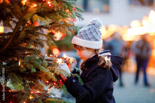 Little cute kid girl having fun on traditional Christmas market during strong snowfall. Happy child enjoying traditional family market in Germany. Schoolgirl standing by illuminated xmas tree. photo