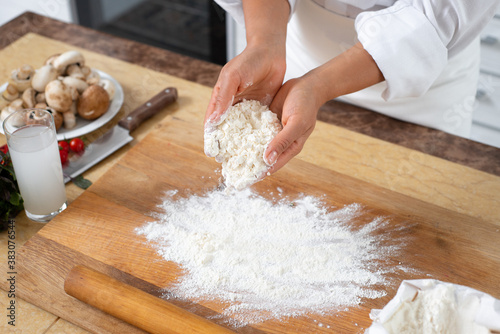 Close-up on the hands of a cook with a handful of white flour. On the table is a wooden board with scattered flour.