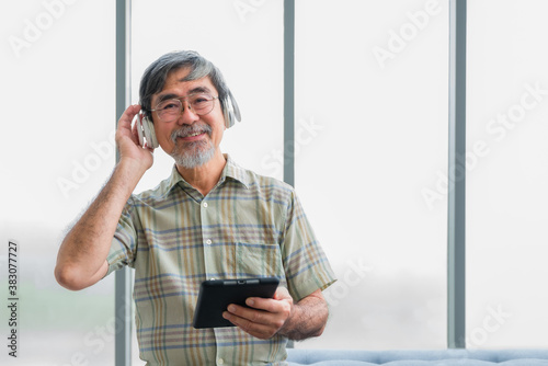 Senior old male wearing white earphone and listening music. Lifestyle after retirement of elderly. Happy asian elderly man sitting on couch wear headphone and sing a song in living room at his home.