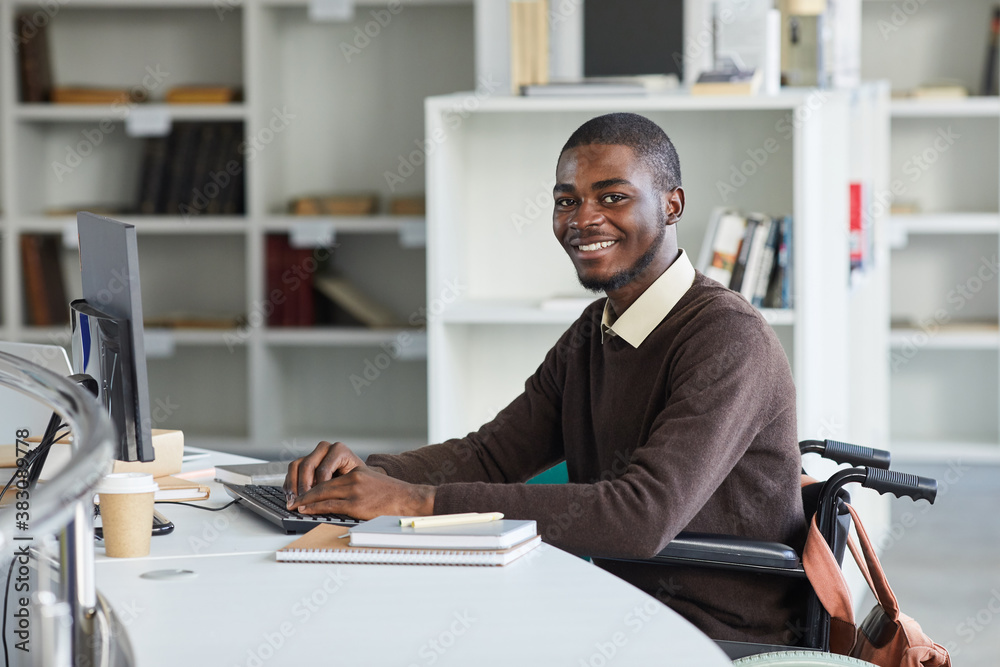 Portrait of disabled African-American man using computer and smiling at camera while studying in college library, copy space Portrait of disabled African-American man using computer and smiling at camera while studying in college library, copy space