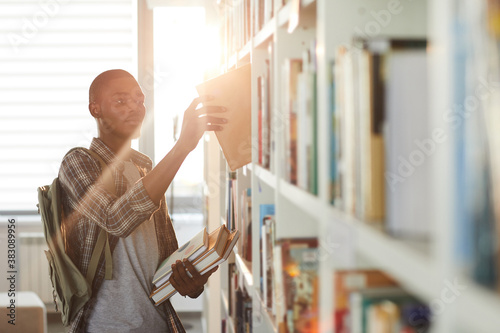 Waist up portrait of young African-American man taking book off shelf in school library against sunlight, copy space