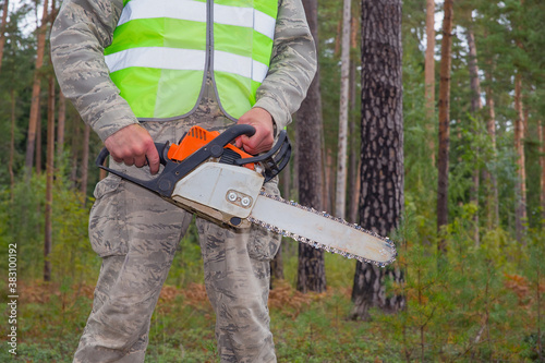 The worker holds a chainsaw in his hands. Close-up photo. In the background there is a pine forest. The concept of cutting forests on the planet. Forestry and timber harvesting.