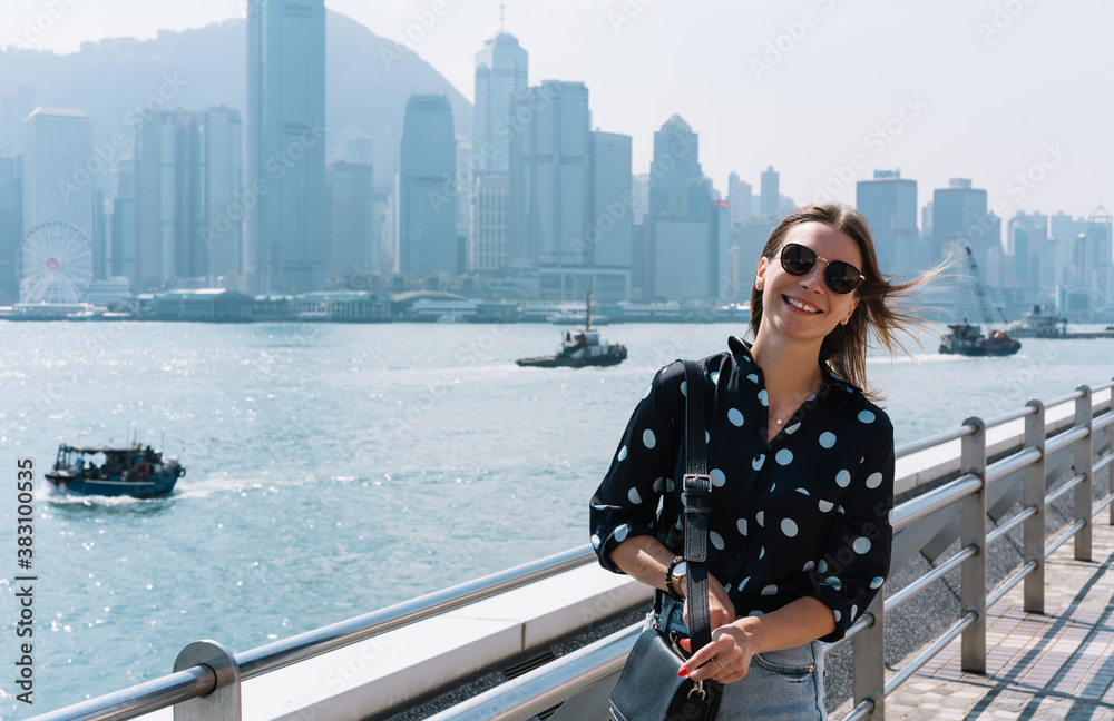 Half length portrait of cheerful caucasian female tourist in sunglasses enjoying vacation explore city on sunny day, positive 20s woman in trendy clothes looking at camera standing on bridge in bay