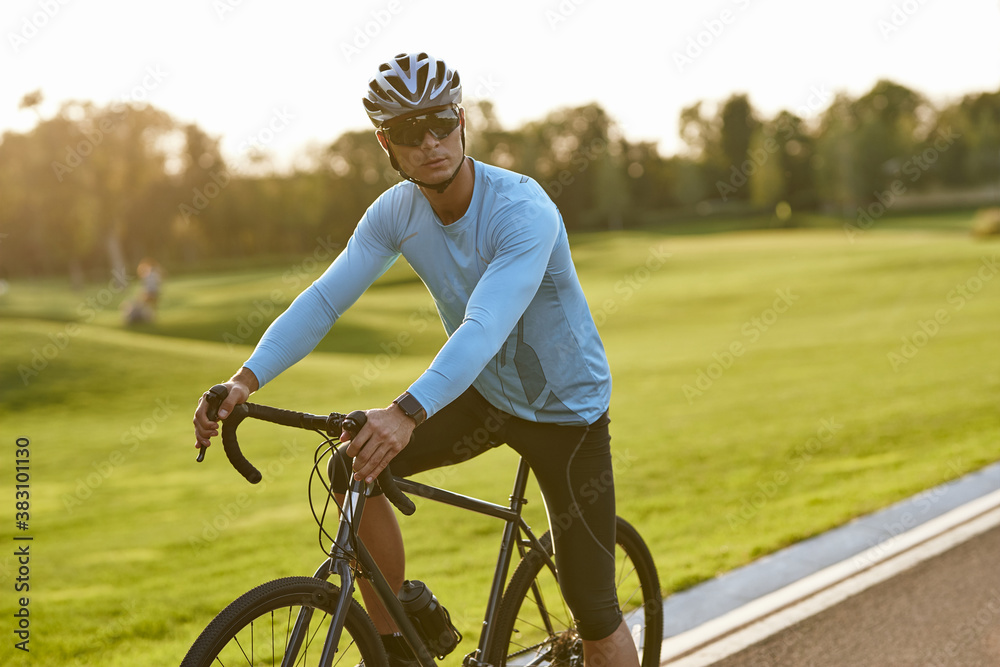 Obraz premium Strong athletic man in sportswear and protective helmet standing with his bicycle on the road in park and looking away, resting after cycling outdoors