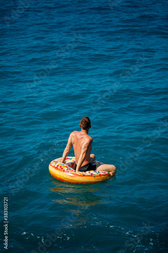A young man floats on an inflatable air ring circle in the sea with blue water. Festive holiday on a happy sunny day. Vacation concept, top view.