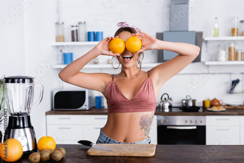 Selective focus of tattooed woman holding oranges near eyes beside cutting board and blender