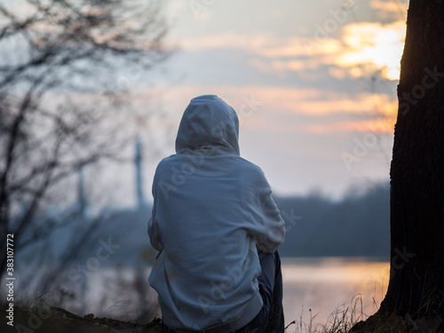 A lonely young man sits on the riverbank at sunset in autumn. Melancholic depressive mood . From the back