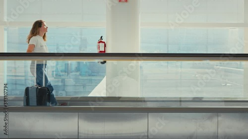 Young woman with suitcase travels on moving walkaway escalator in empty airport terminal. Light and bright ambience of new built airport