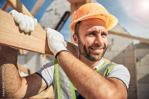 Positive delighted young constructor carrying wooden beam