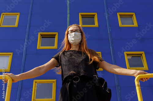 young redhead girl in protective mask against COVID virus, looks worried at the side in front of blue wall with yellow windows. Coronavirus, health protection, quarantine, safety and pandemic concept