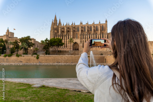 Young light-skinned girl taking a picture of the cathedral of Palma de Mallorca with her phone on her holidays.