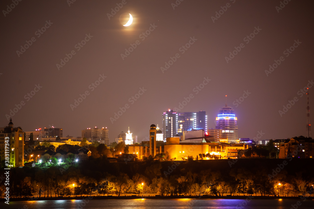 Above the night city shines the moon against the yellow gray sky