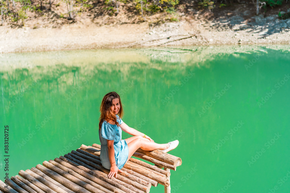 Beautiful girl with long hair in a dress on a bridge above a mountain lake with clear water and a view of a green forest. Copy space