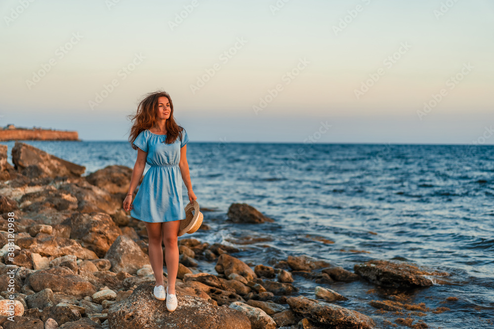 A young beautiful woman in a dress with a hat stands on the beach at sunset and looks at the setting sun, Lonely girl dreams of love.