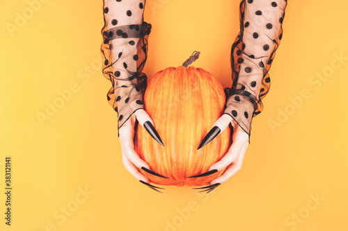 Halloween concept. Witch hands with long black nails are holding a pumpkin on the orange background. Flat-lay, top view.