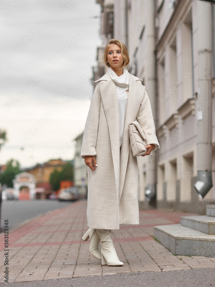 Elegant woman in white dress, hessian boots and coat walking at city street. Fall autumn fashion look. Pretty tall stylish young gitl with fashionable makeup and hair style. Elegant lady. Full length