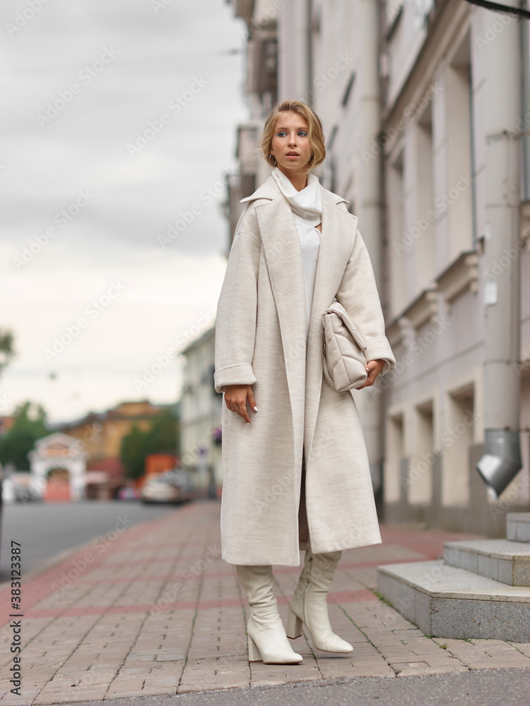 Fototapeta premium Elegant woman in white dress, hessian boots and coat walking at city street. Fall autumn fashion look. Pretty tall stylish young gitl with fashionable makeup and hair style. Elegant lady. Full length