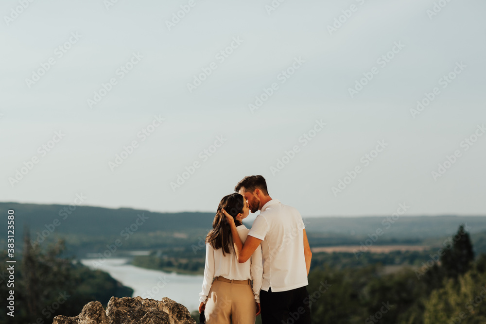 Back view of a young stylish couple enjoying life and nature. Man embracing and kissing woman. Panorama of the mountains.