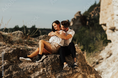 Happy couple are sitting on a big rock while enjoying time together. Young beautiful romantic couple in stylish clothes sitting on a stone on the background of cliff and trees. © eduard
