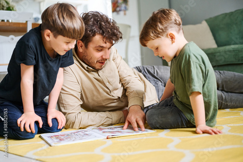 Father with sons reading book, spending time together at home and lying on the floor