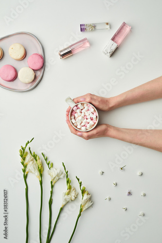 Minimal background composition of female hands holding cup of sweet cocoa over while table background with floral decor and pink accents, copy space