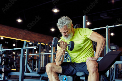 Getting strong arms. Focused mature man in sportswear lifting heavy dumbbells and pumping his biceps while working out at gym photo