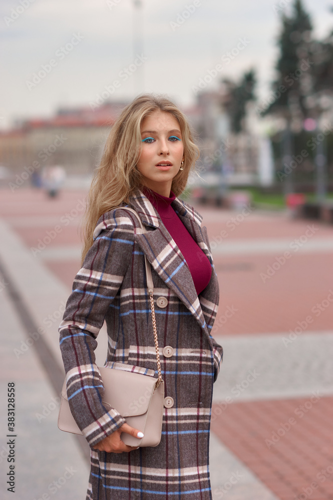 Fototapeta premium Elegant woman walking city street. Fashion fall autumn look. Pretty caucasian blonde stylish girl wearing blue jeans, wine red pullover and squared wool coat. Female with makeup and wavy blonde hair