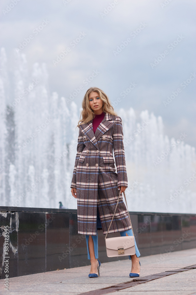 Elegant woman walking city street. Fashion fall autumn look. Pretty caucasian blonde stylish girl wearing blue jeans, wine red pullover and squared wool coat. Female with makeup and wavy blonde hair Elegant woman walking city street. Fashion fall autumn look. Pretty caucasian blonde stylish girl wearing blue jeans, wine red pullover and squared wool coat. Female with makeup and wavy blonde hair